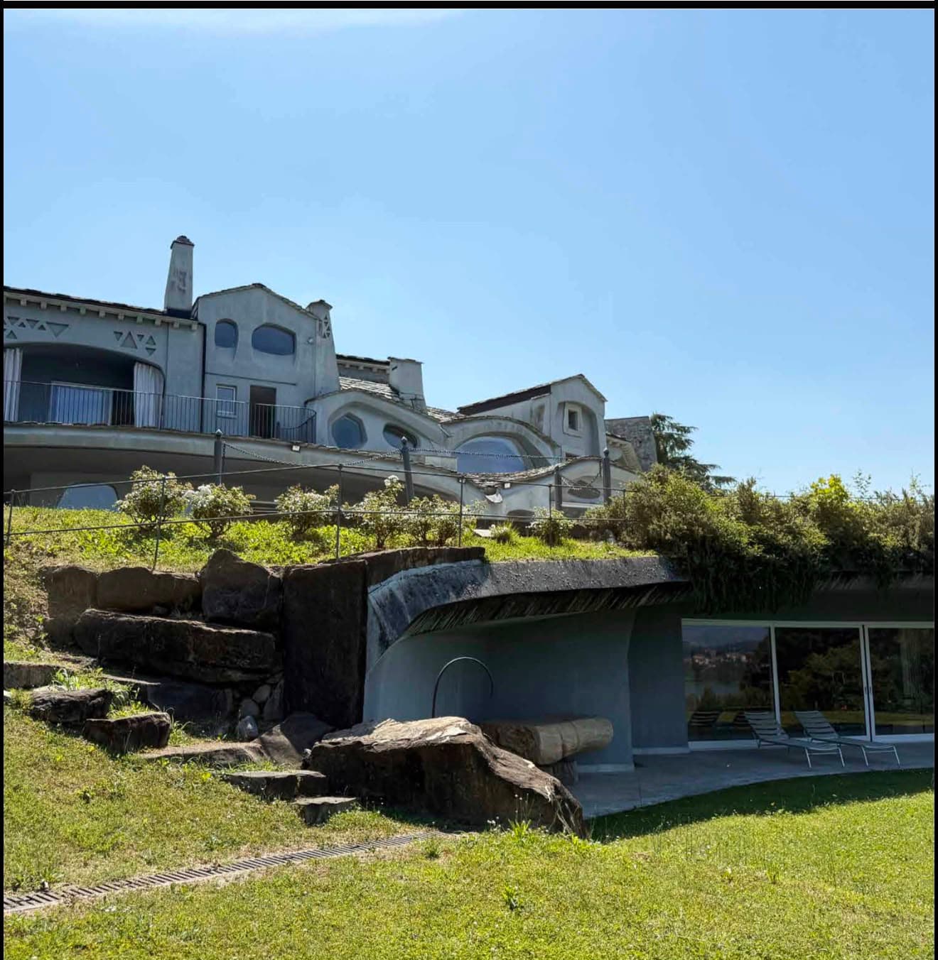 Villa grigia dalle forme curve e ampie vetrate su una collina verdeggiante e soleggiata.