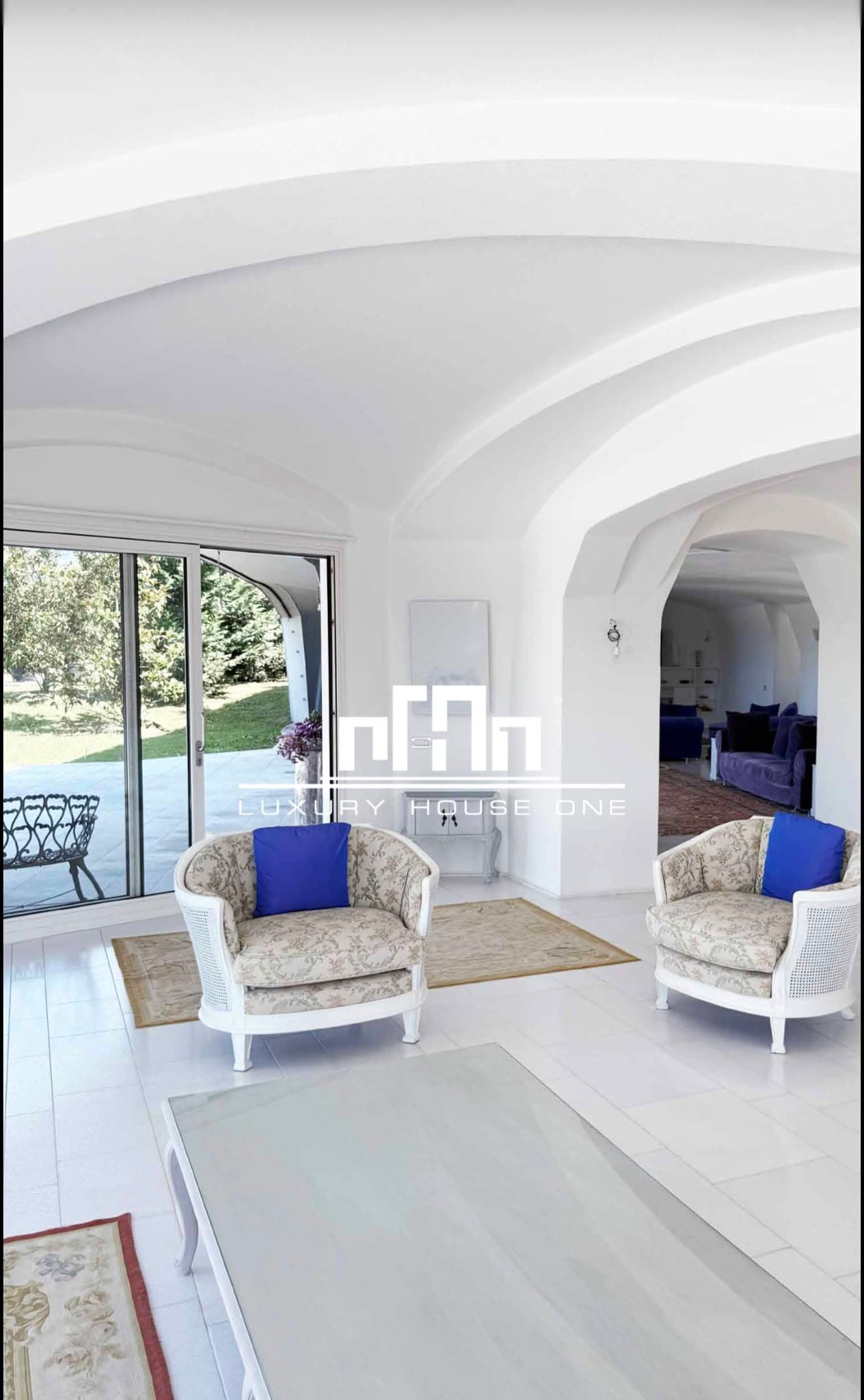 Soggiorno bianco con soffitto a volta, poltrone fiorite e vista panoramica sul giardino esterno.